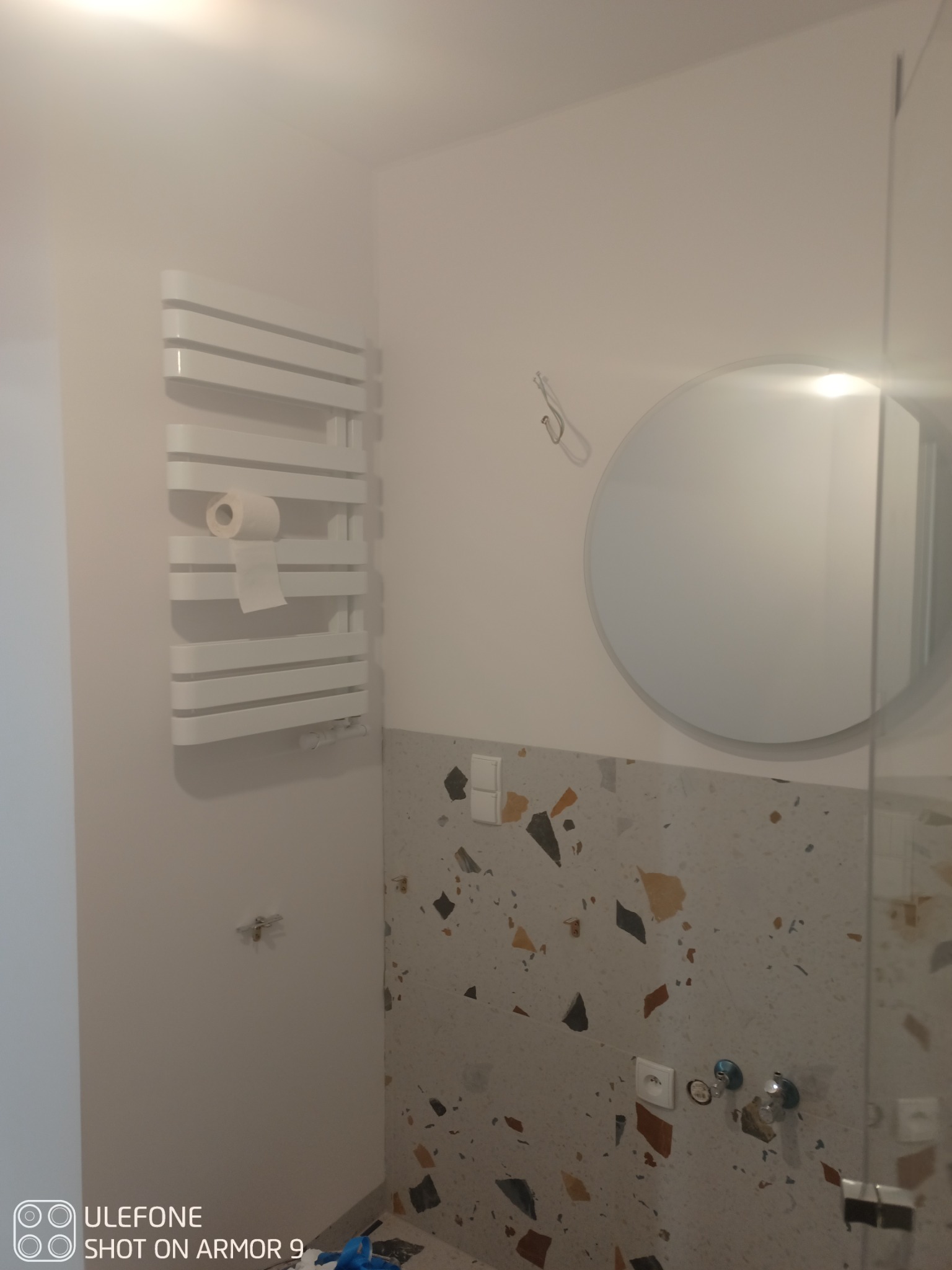 Biała łazienka w trakcie remontu: widoczny grzejnik drabinkowy z rolką papieru, okrągłe lustro, ściana wyłożona płytkami w stylu lastryko oraz wystające kable i rury.