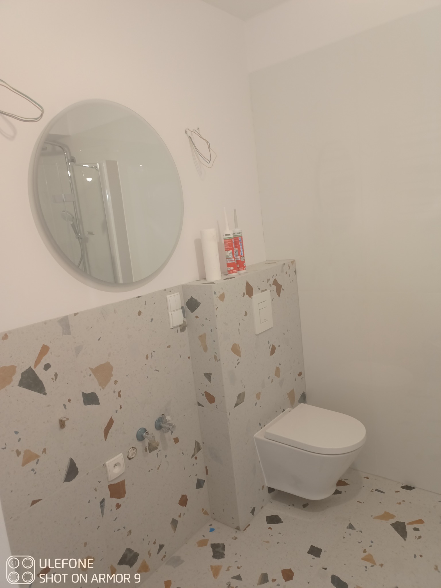 Nowoczesna toaleta podwieszana z zabudową z płytek lastryko, widoczne dwa kartusze z silikonem sanitarnym, okrągłe lustro z odbiciem kabiny prysznicowej oraz wystające przewody elektryczne.