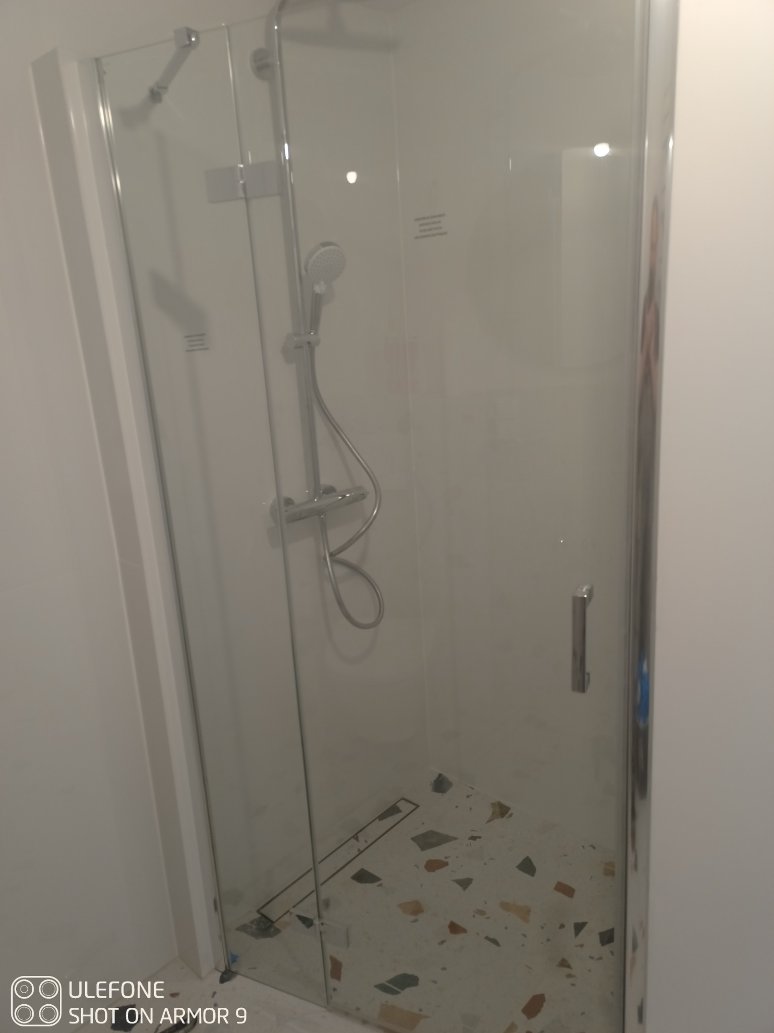 Kabina prysznicowa z chromowanymi detalami i wzorzystą posadzką z terazzo, widok z zewnątrz, zamknięte drzwi.