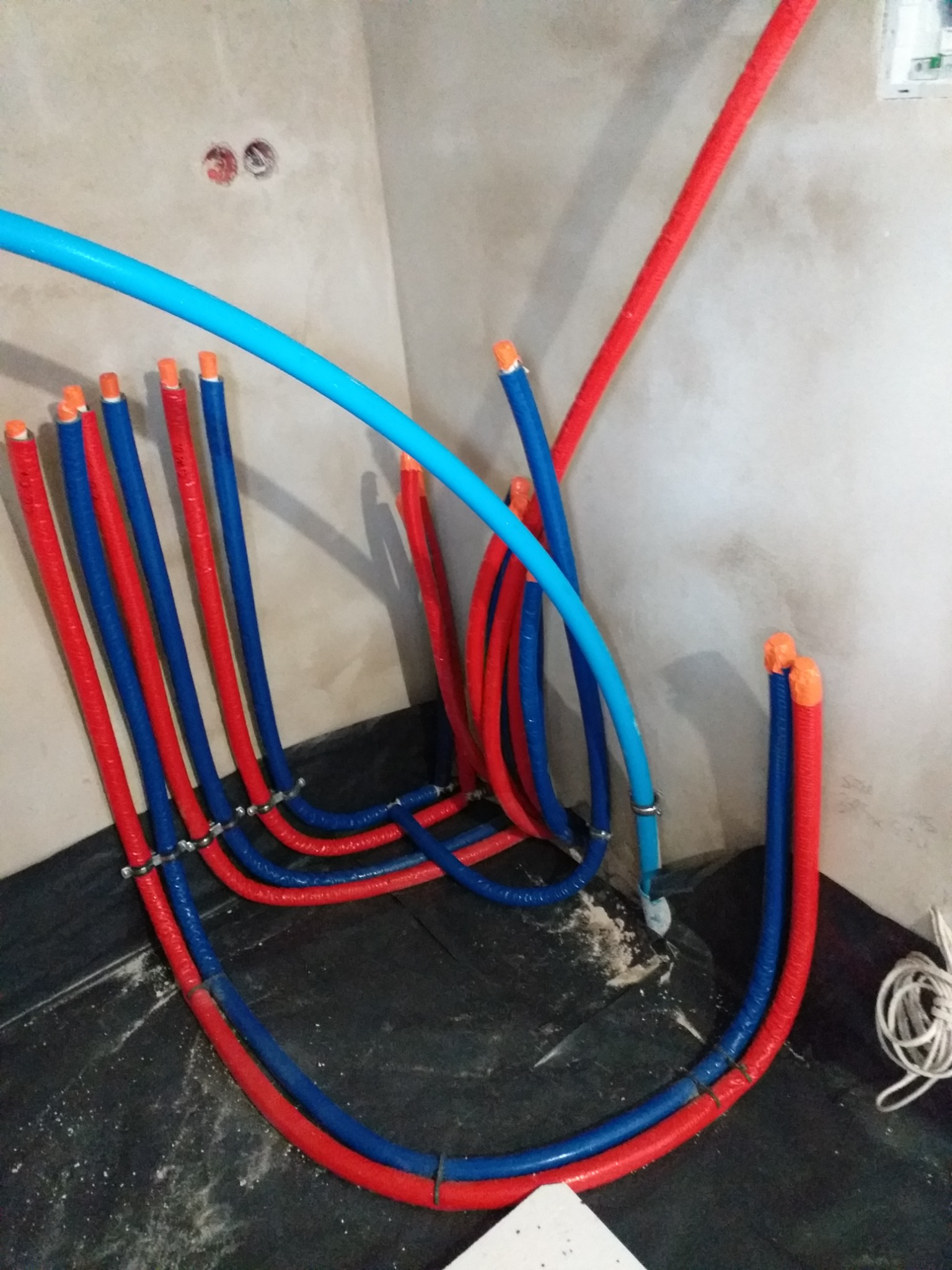 Instalacja wodna w trakcie montażu: rury w izolacji czerwonej i niebieskiej, z pomarańczowymi zaślepkami, prowadzone wzdłuż ściany i podłogi wyłożonej folią ochronną.