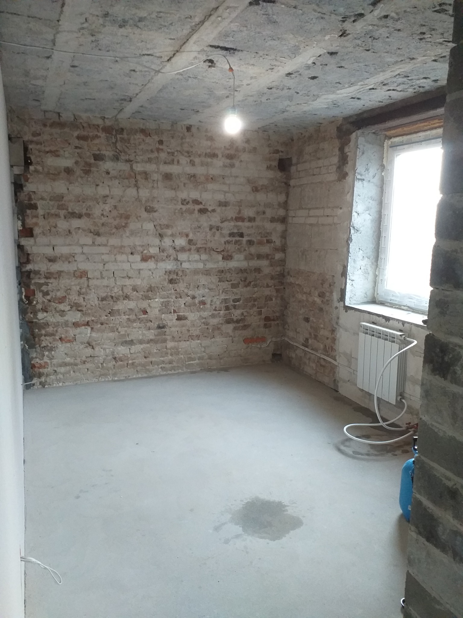 Surowy pokój z odsłoniętą cegłą i betonowym sufitem przed remontem, widoczny grzejnik pod oknem i ślady wilgoci na podłodze.