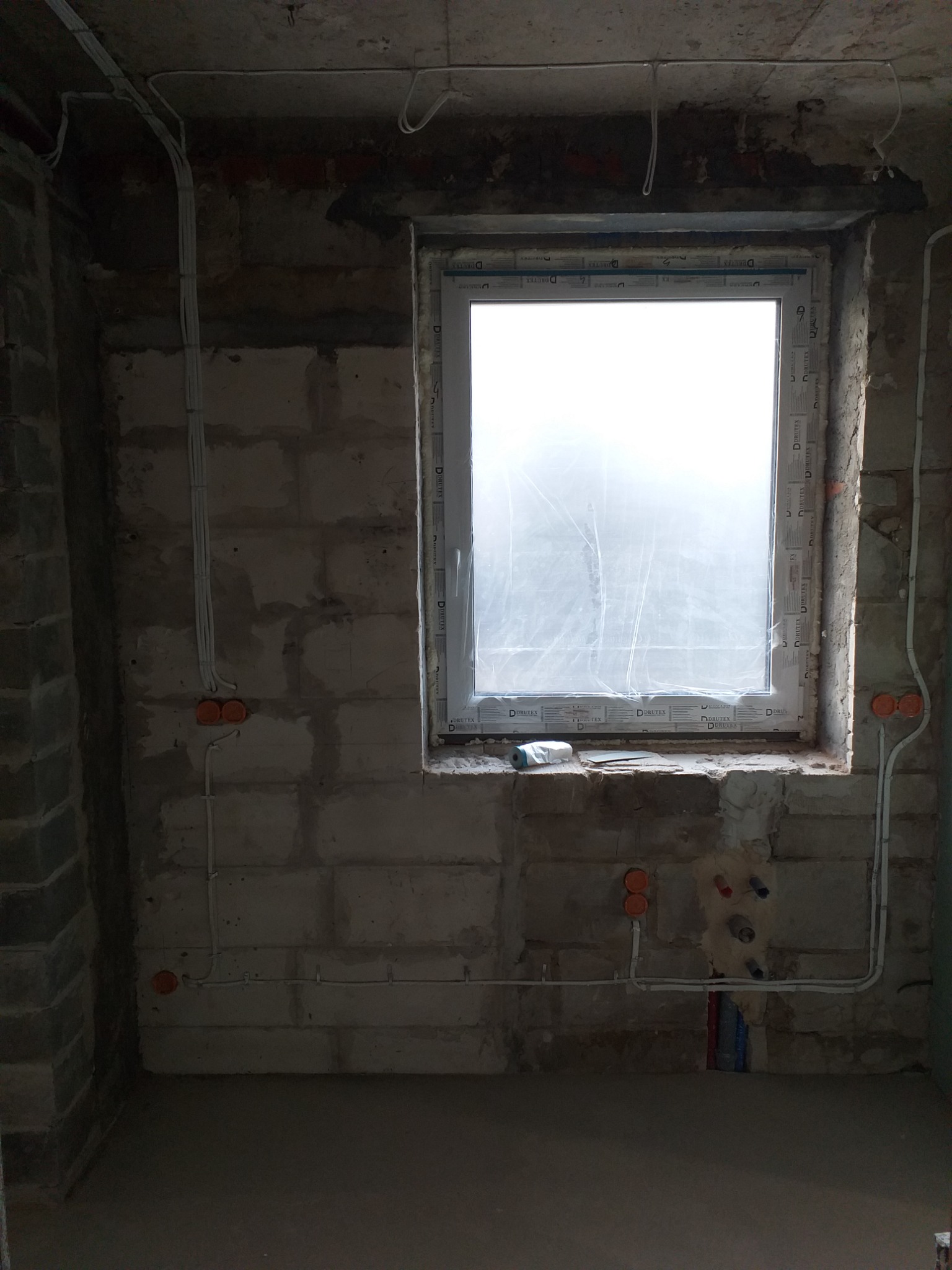 Surowy stan wnętrza w trakcie prac remontowych: widoczne przewody elektryczne w peszlach, puszki instalacyjne na ścianach z betonu komórkowego oraz okno zabezpieczone folią malarską.
