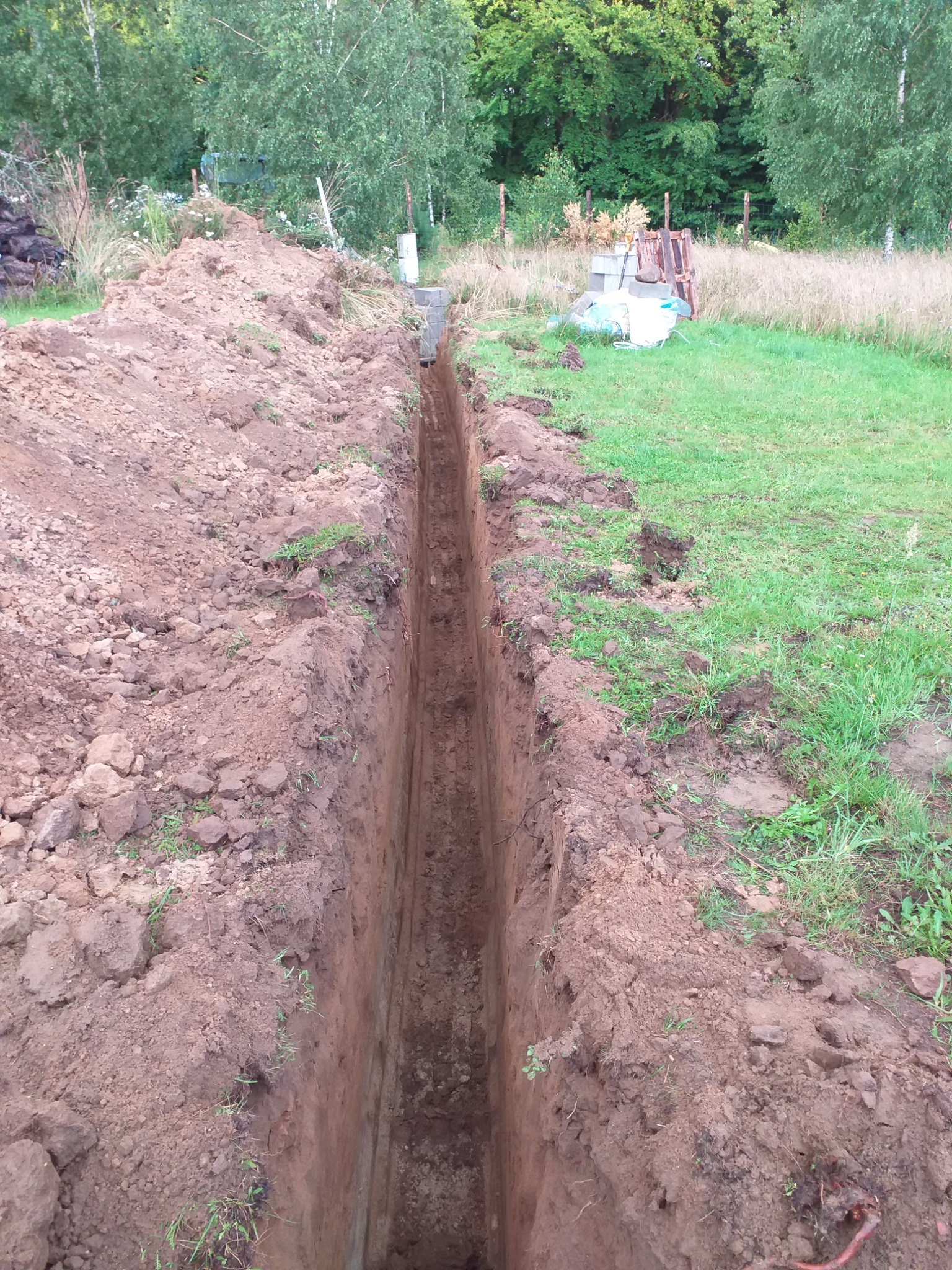 Wąski wykop w ziemi z równymi ścianami, przygotowany pod instalację, z widocznymi betonowymi elementami na jednym końcu, otoczony trawą i drzewami.