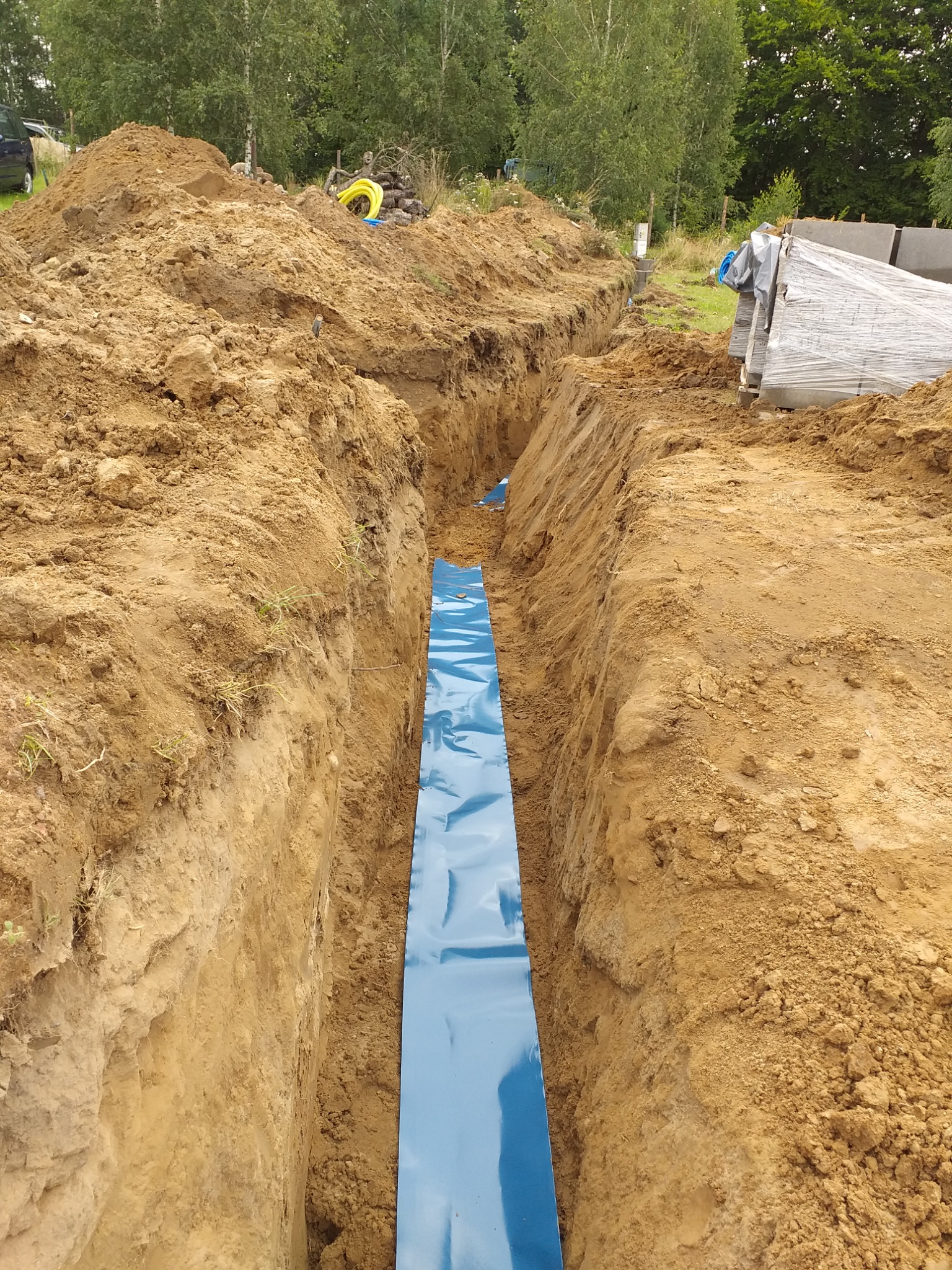 Wykopany rów z niebieską folią izolacyjną na dnie, przygotowany do instalacji podziemnej, z widoczną stertą ziemi i elementami instalacji na powierzchni.