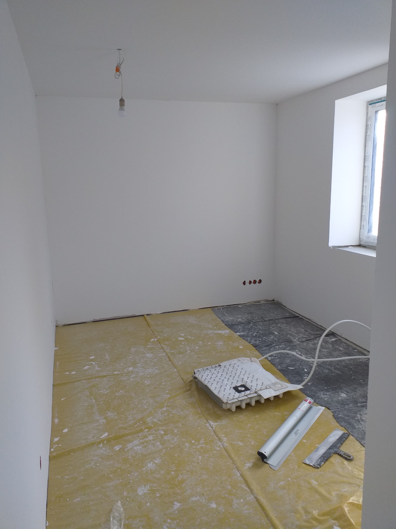 Pokój w trakcie przygotowania do malowania: zabezpieczona folią podłoga, widoczne narzędzia malarskie, gładkie, białe ściany, surowe gniazdka elektryczne, okno z widocznymi uszczelkami.