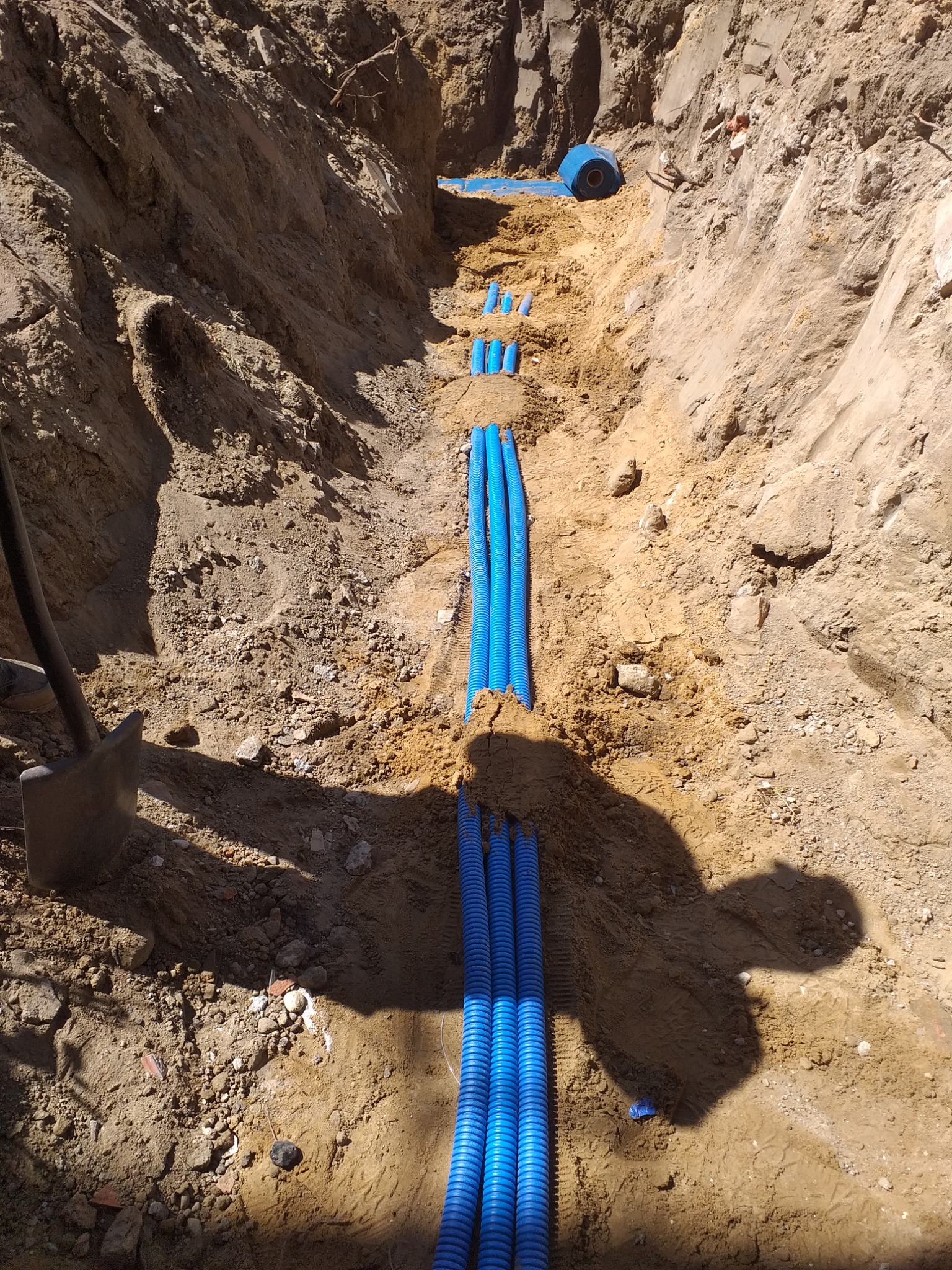 Wykop z ułożonymi niebieskimi rurami karbowanymi, widoczna łopata i rolka niebieskiej folii zabezpieczającej.