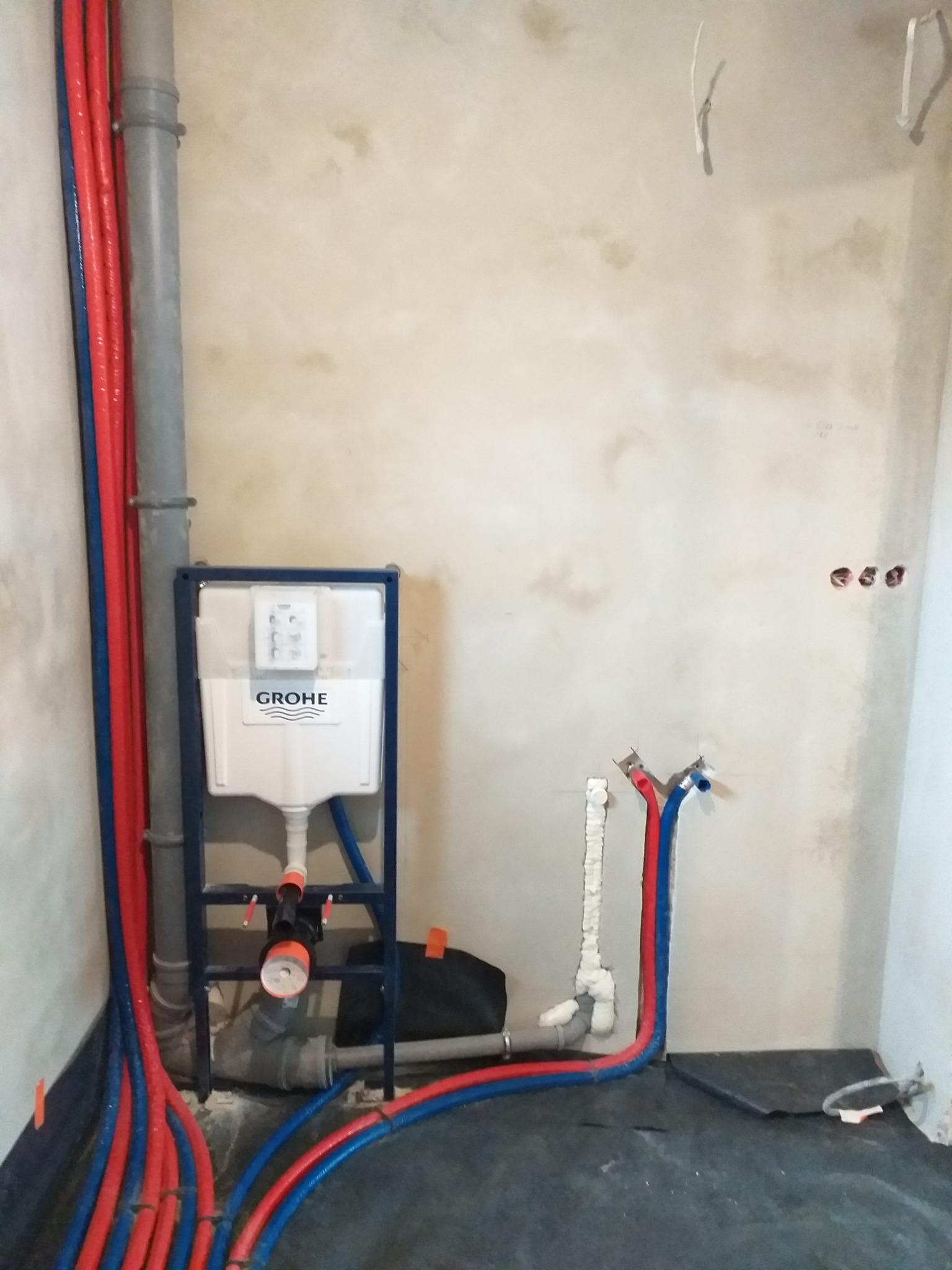 Stelaż podtynkowy Grohe z podłączonymi rurami wodnymi w kolorze czerwonym i niebieskim oraz szarą rurą kanalizacyjną, widoczne otwory w ścianie na kable elektryczne, surowa ściana z widocznymi...