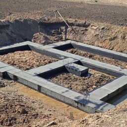 USŁUGI BUDOWLANE BUD-AR Arkadiusz Mamach - Wylane betonowe ławy fundamentowe z wystającymi prętami zbrojeniowymi, zabezpieczone czarną izolacją przeciwwilgociową, w wykopie ziemnym z kałużami wody.