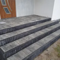 Schody wejściowe do domu wyłożone szarą kostką brukową z czarnymi obrzeżami, prowadzące do drzwi wejściowych z szybami i białej elewacji.