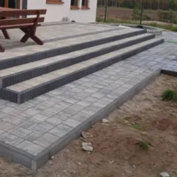 Ułożona kostka brukowa tworząca taras z trzema stopniami i chodnikiem, obrzeża z ciemnej kostki, w tle budynek mieszkalny i teren zielony.