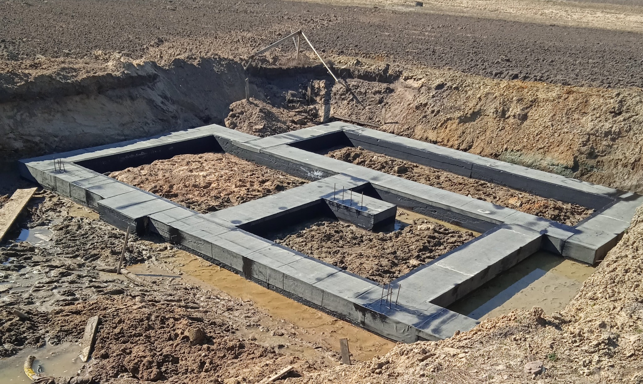 Wylane betonowe ławy fundamentowe z wystającymi prętami zbrojeniowymi, zabezpieczone czarną izolacją przeciwwilgociową, w wykopie ziemnym z kałużami wody.