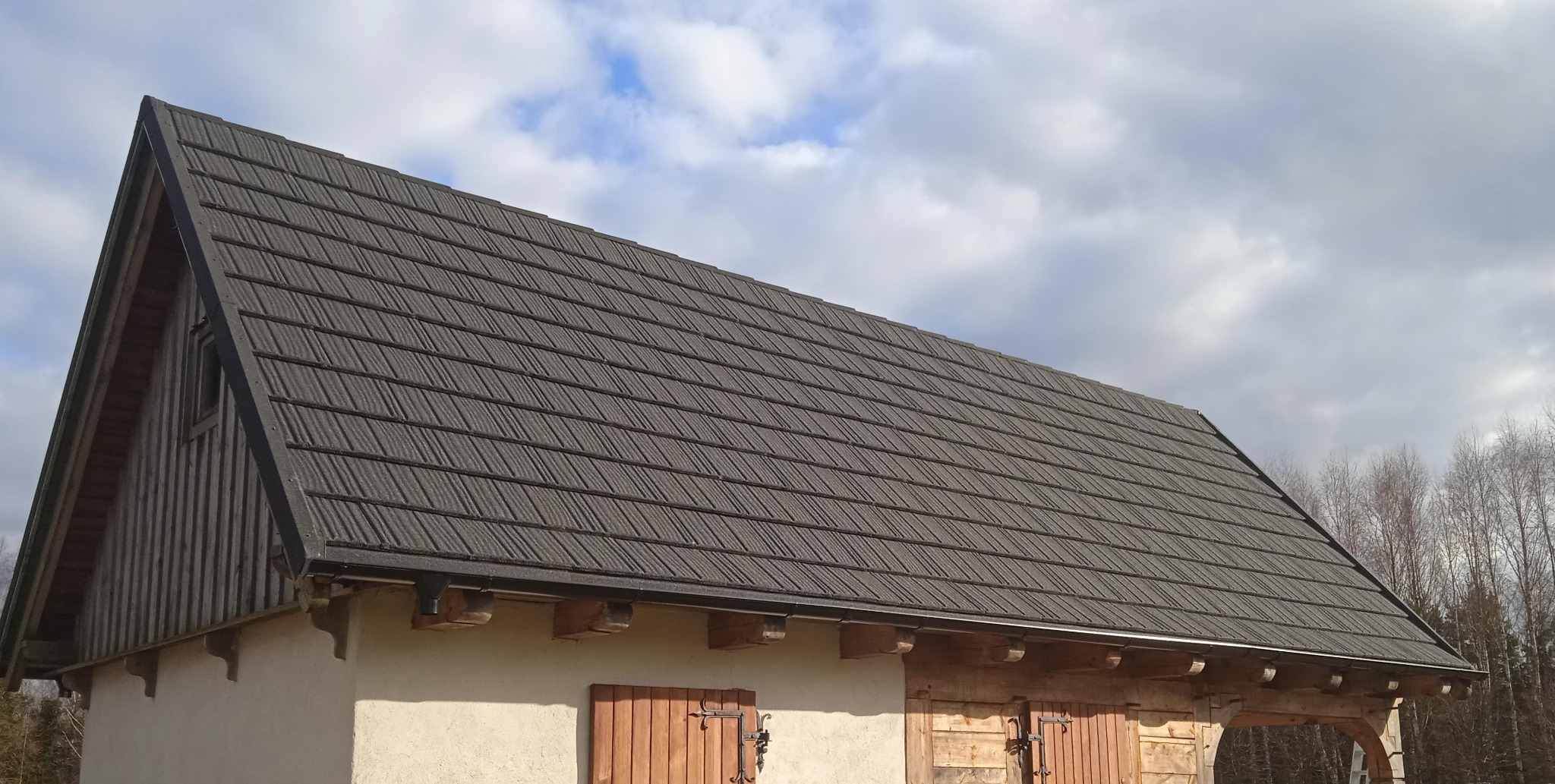 Dom z ciemnoszarą, modułową dachówką, drewnianą szalówką na szczycie i okiennicami, na tle pochmurnego nieba.