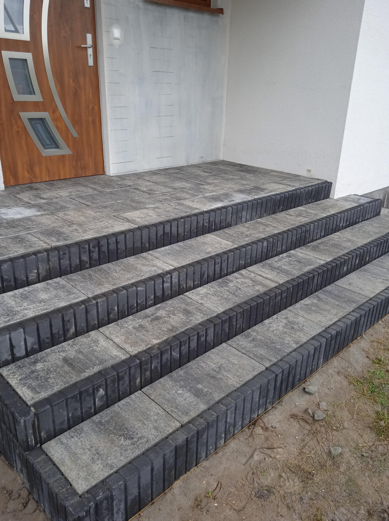 Schody wejściowe do domu wyłożone szarą kostką brukową z czarnymi obrzeżami, prowadzące do drzwi wejściowych z szybami i białej elewacji.