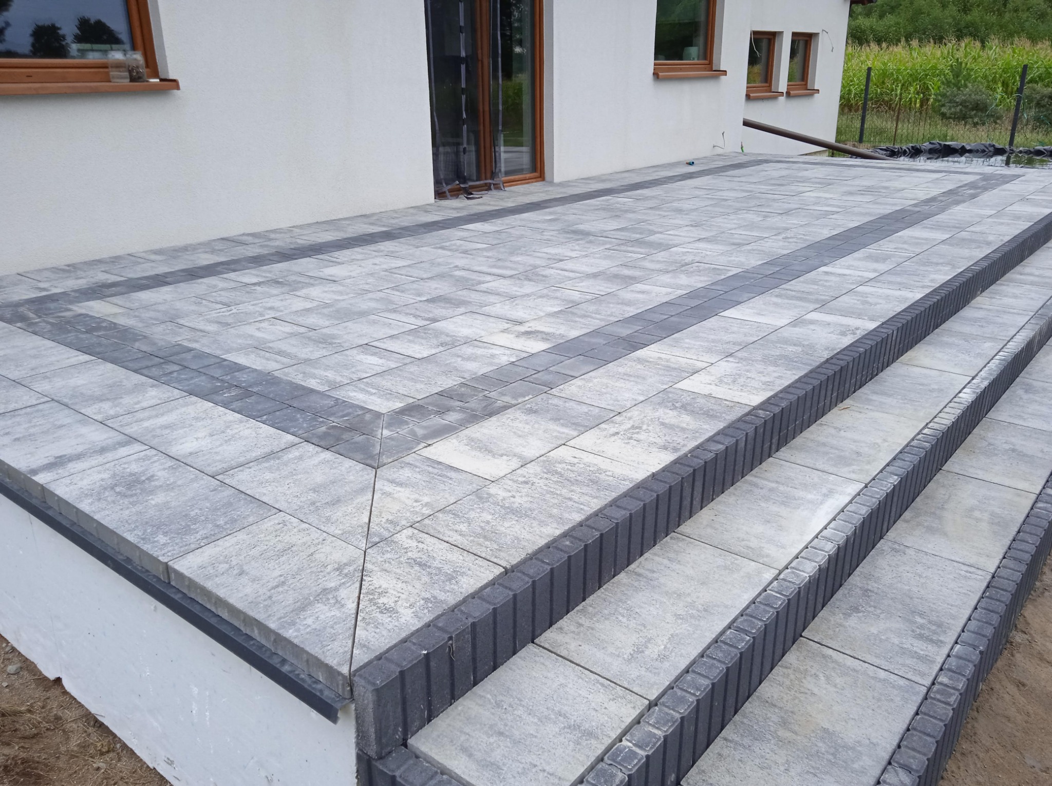 Wykończony taras z kostki brukowej w odcieniach szarości, z subtelnym wzorem i schodami, przylegający do białego budynku z brązowymi ramami okiennymi.