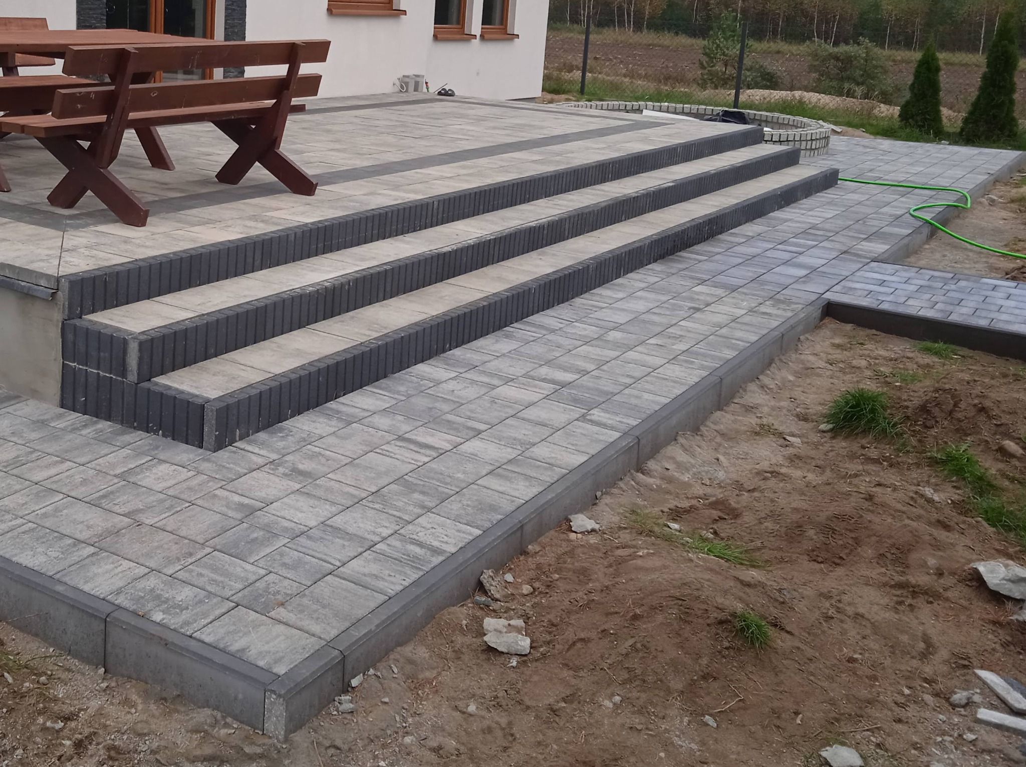 Ułożona kostka brukowa tworząca taras z trzema stopniami i chodnikiem, obrzeża z ciemnej kostki, w tle budynek mieszkalny i teren zielony.