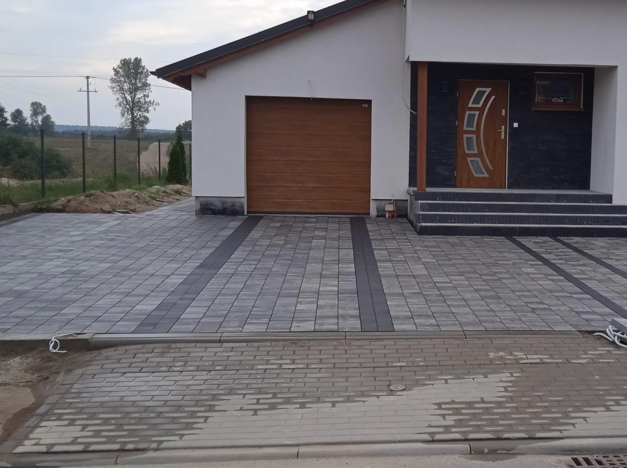 Nowoczesny dom z szarą kostką brukową na podjeździe, ułożoną w geometryczny wzór z ciemniejszymi pasami. Widoczny front domu z brązową bramą garażową i drzwiami wejściowymi z elementami...