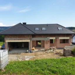 Surowy stan domu jednorodzinnego z czerwonej cegły z czarnym dachem, widoczne otwory okienne i garażowe, na pierwszym planie zbiornik na wodę i teren budowy z drewnem.