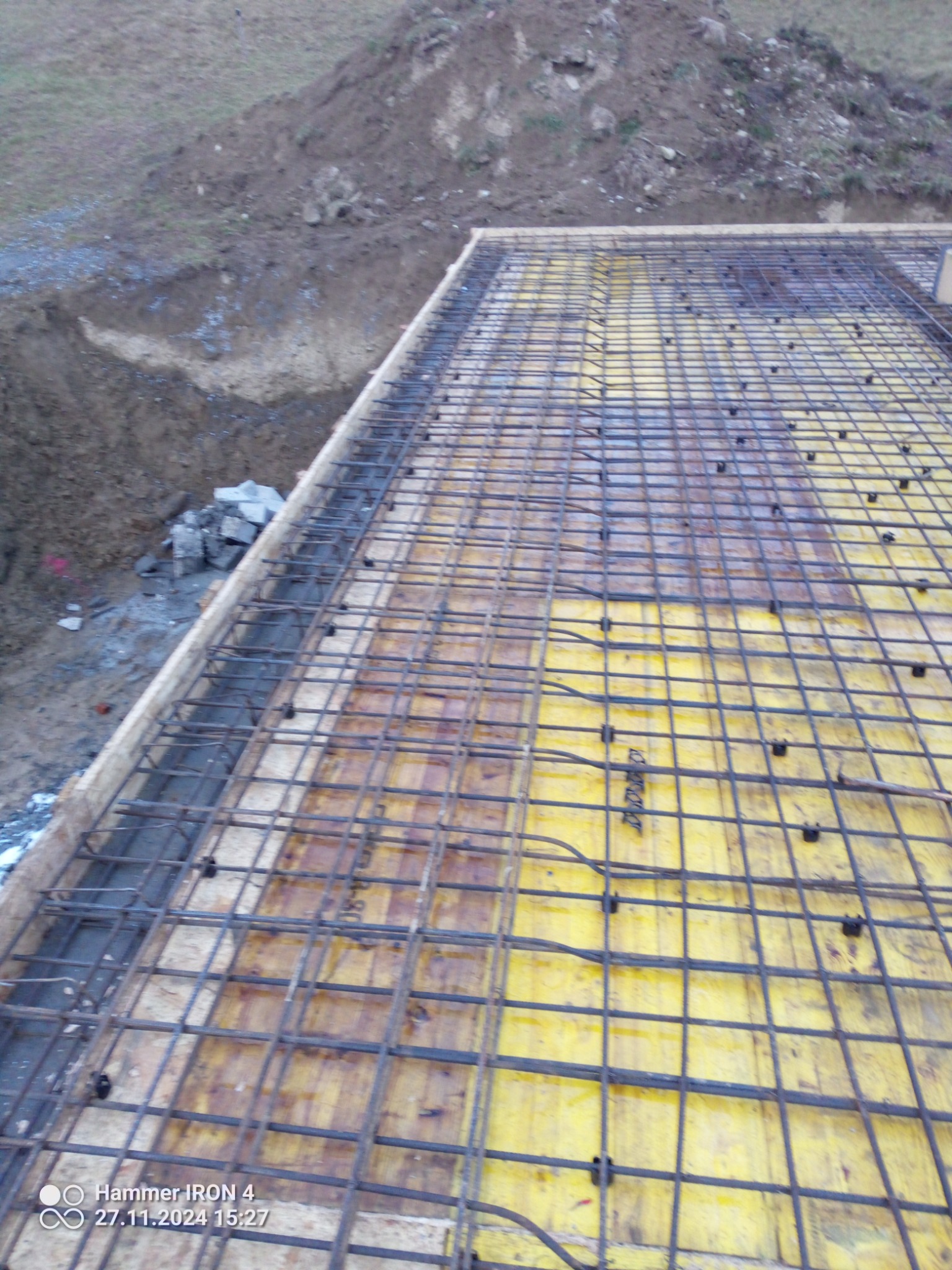Zbrojenie fundamentu z siatki stalowej na żółtych płytach szalunkowych, widok z góry, obok wykop z ziemią i kamieniami.