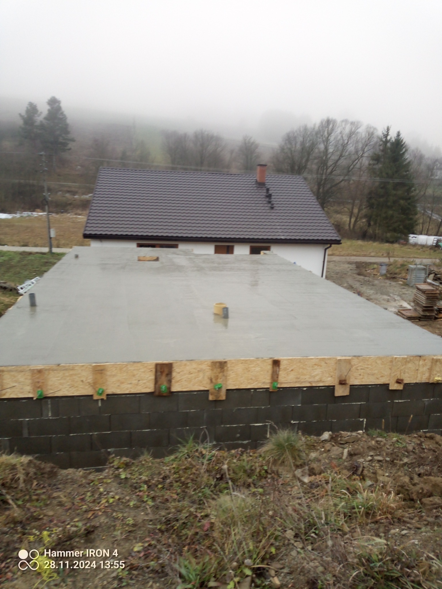 Wylany betonowy fundament z szalunkiem, widoczny fragment domu w tle, pochmurna pogoda.