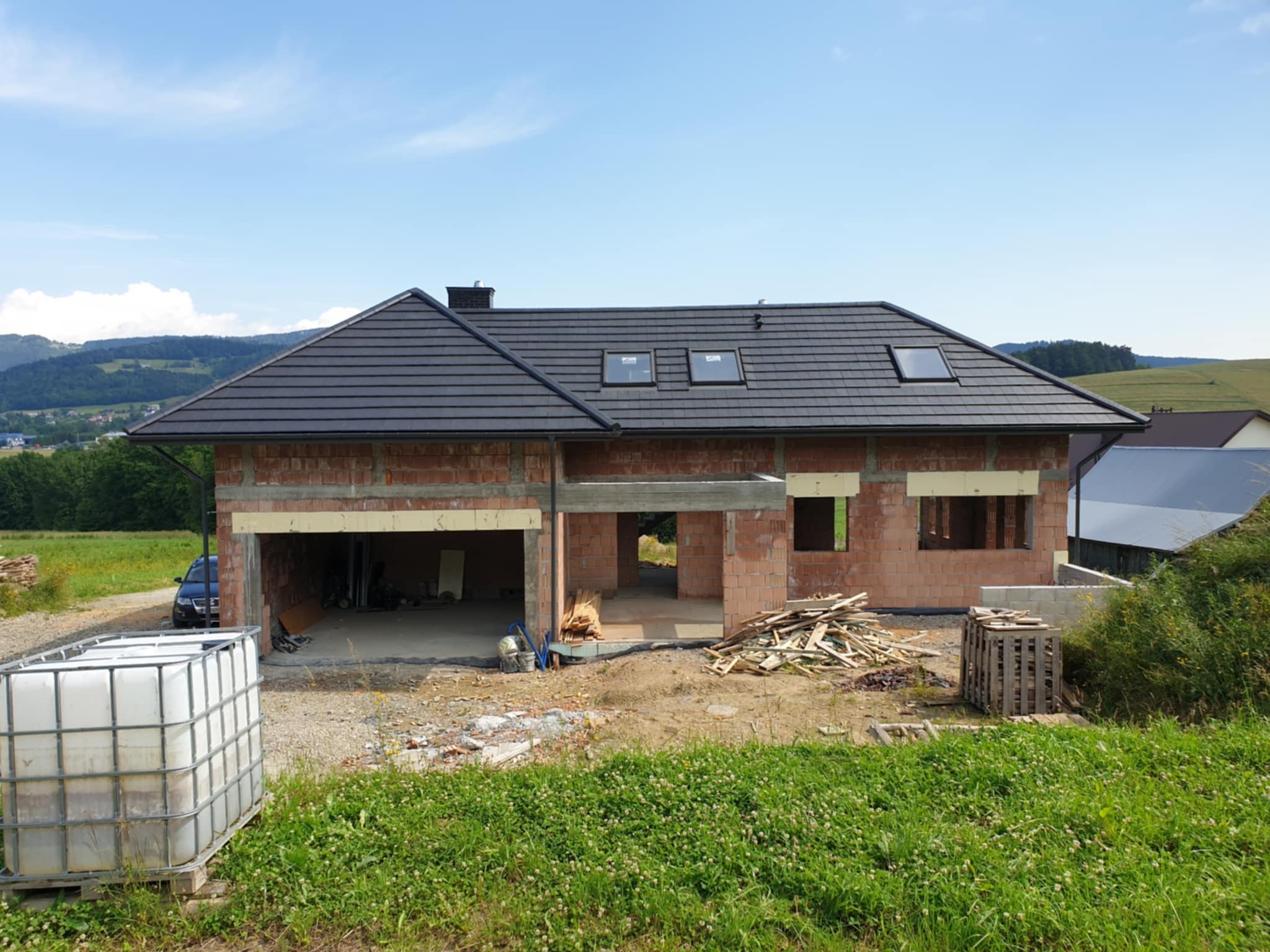 Surowy stan domu jednorodzinnego z czerwonej cegły z czarnym dachem, widoczne otwory okienne i garażowe, na pierwszym planie zbiornik na wodę i teren budowy z drewnem.