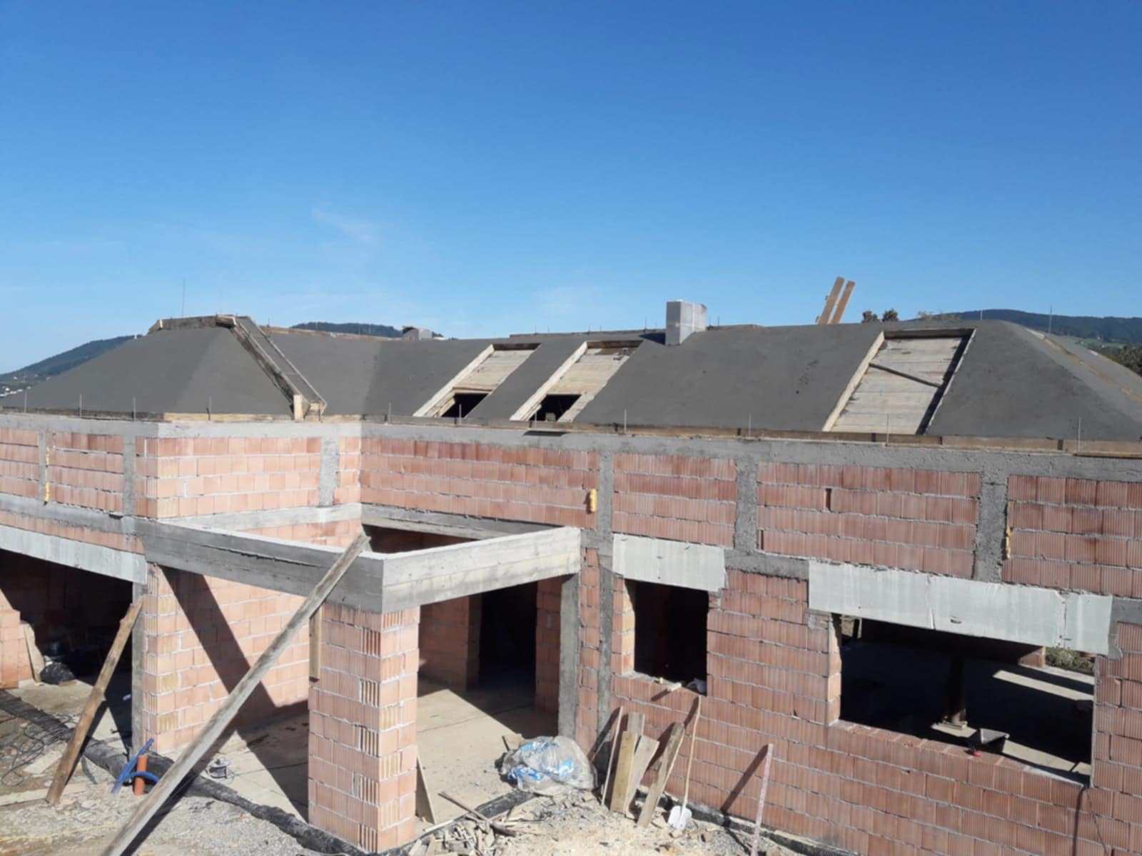 Wznoszenie ścian parterowego domu z czerwonej cegły, widoczne otwory okienne i drzwiowe, w tle konstrukcja dachu z deskowaniem i pokryciem papą oraz zielone wzgórza.