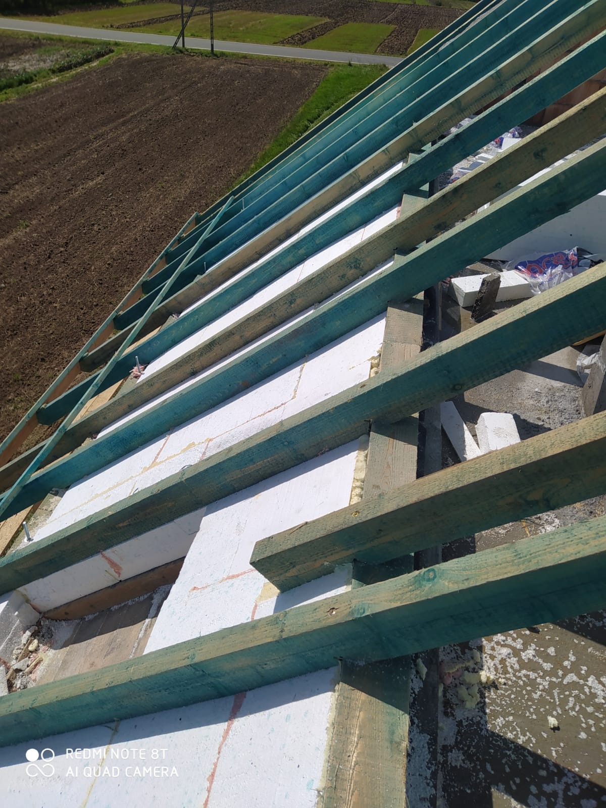 Wnętrze konstrukcji dachu z widoczną więźbą dachową zaimpregnowaną na zielono i izolacją ze styropianu, z widokiem na pole w tle.