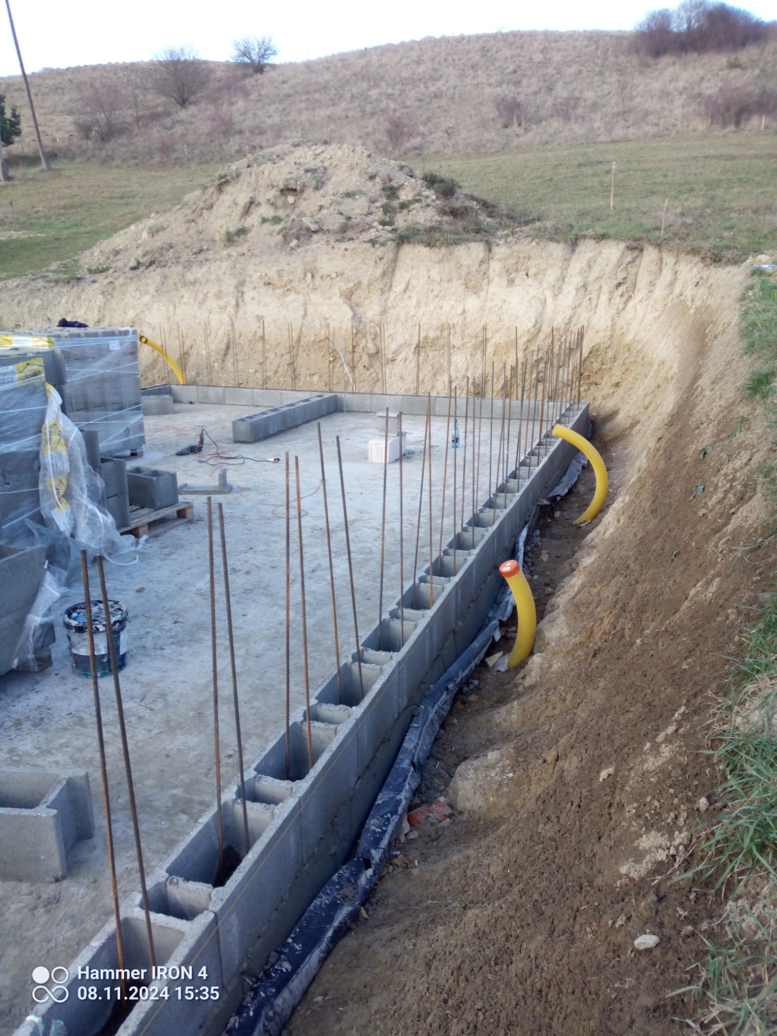 Wznoszenie fundamentów z bloczków betonowych z wystającymi prętami zbrojeniowymi, widoczne żółte rury instalacyjne w ziemi i wykop ziemny pod budowę.