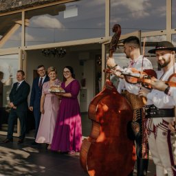 Summer Band - Zespół w strojach ludowych gra na skrzypcach i kontrabasie podczas powitania gości weselnych przed budynkiem z przeszklonymi drzwiami.