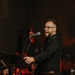 Summer Band - Wokalista z saksofonem śpiewa do mikrofonu podczas występu zespołu muzycznego z Nowego Sącza. Drugi muzyk gra na gitarze w tle, scena oświetlona czerwonym światłem.