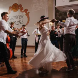 Zespół muzyczny grający na weselu, para młoda tańczy na parkiecie, w tle goście i dekoracja sali weselnej z napisem 'Dworek Jeziorki'.