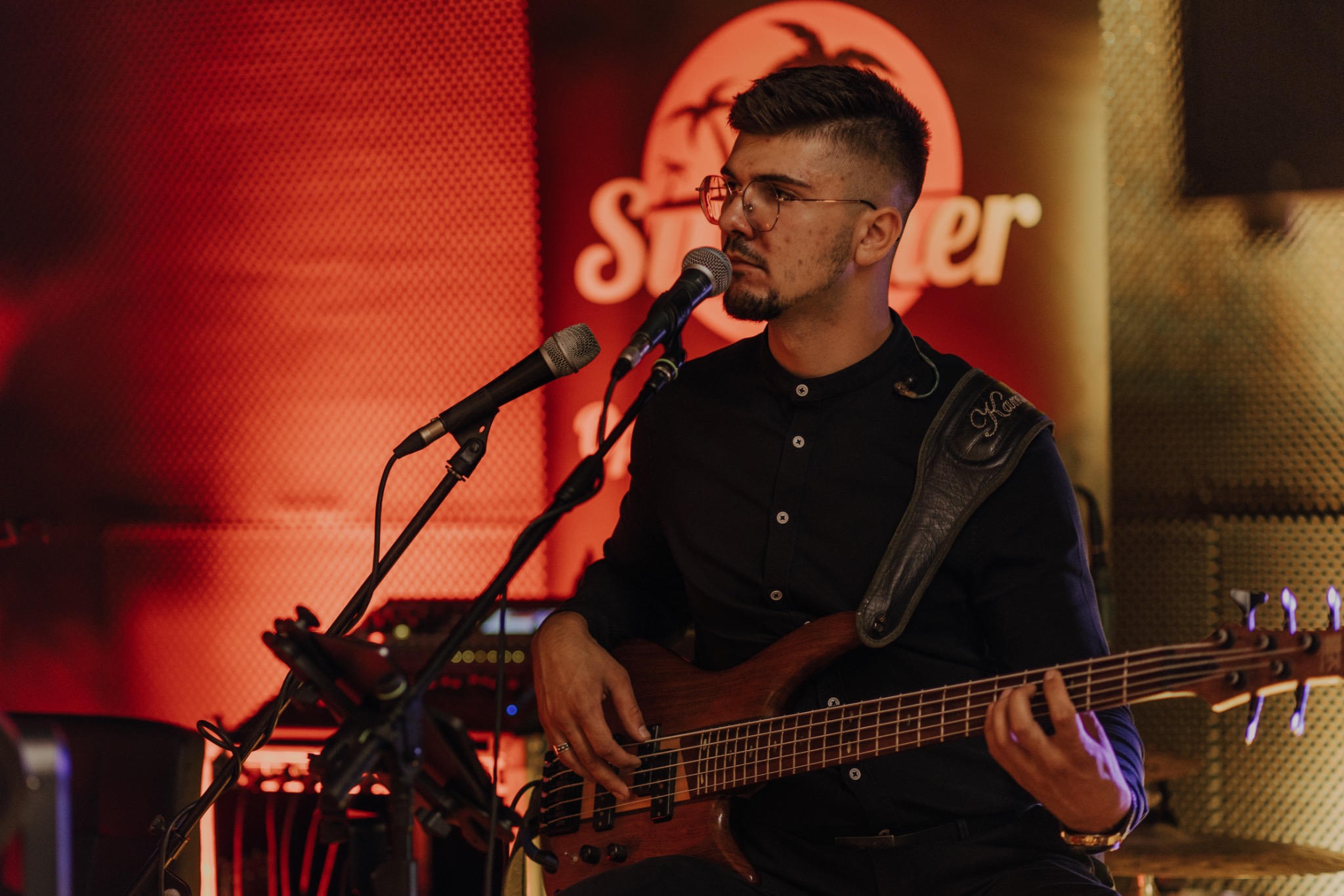 Basista w okularach gra na basie na scenie z czerwonym oświetleniem i mikrofonami. Widoczny logotyp na pasku.
