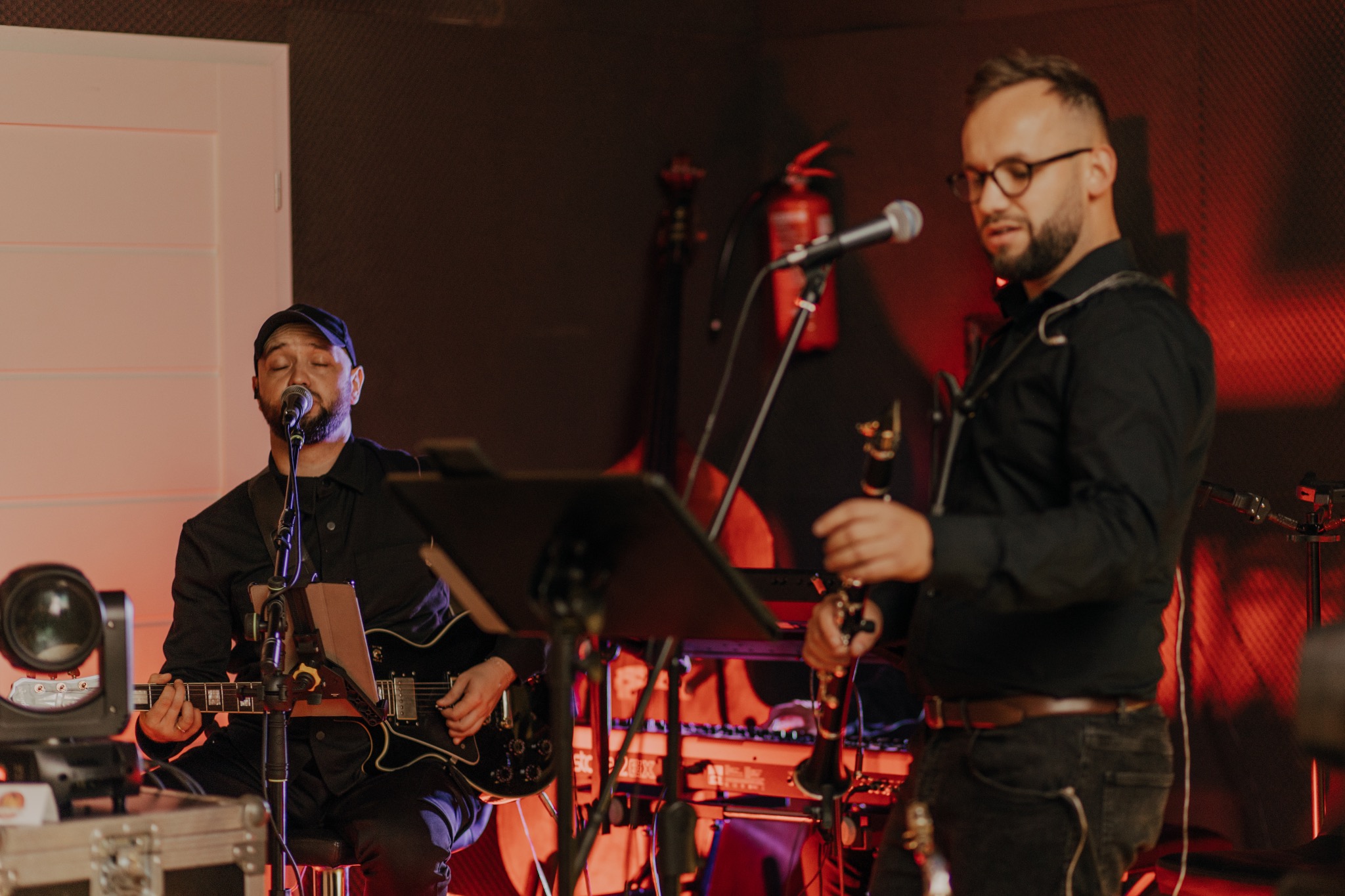 Zespół muzyczny w trakcie występu: gitarzysta śpiewa, klarnecista gotowy do gry, czerwone oświetlenie sceniczne, widoczny syntezator i gaśnica w tle.