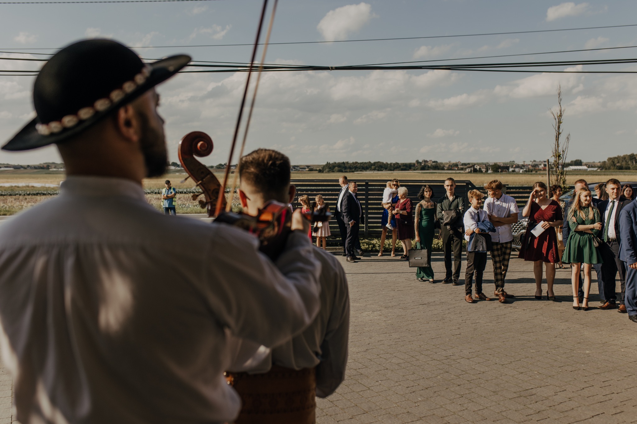 Muzyk w tradycyjnym stroju gra na skrzypcach dla gości weselnych. Ujęcie zza pleców muzyka, widoczni goście w tle i krajobraz. Słoneczny dzień.