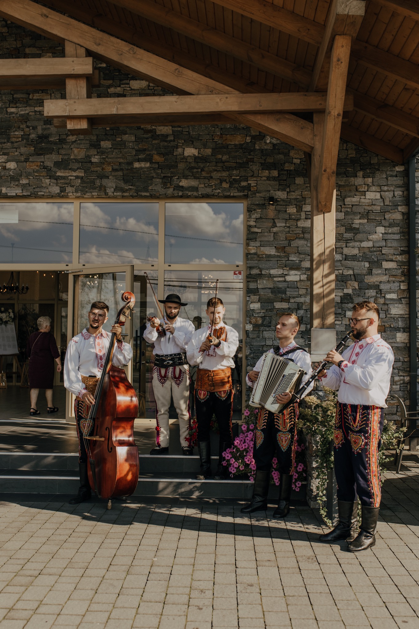 Zespół muzyczny w strojach ludowych grający na instrumentach: kontrabas, skrzypce, akordeon i klarnet przed budynkiem z kamienia.