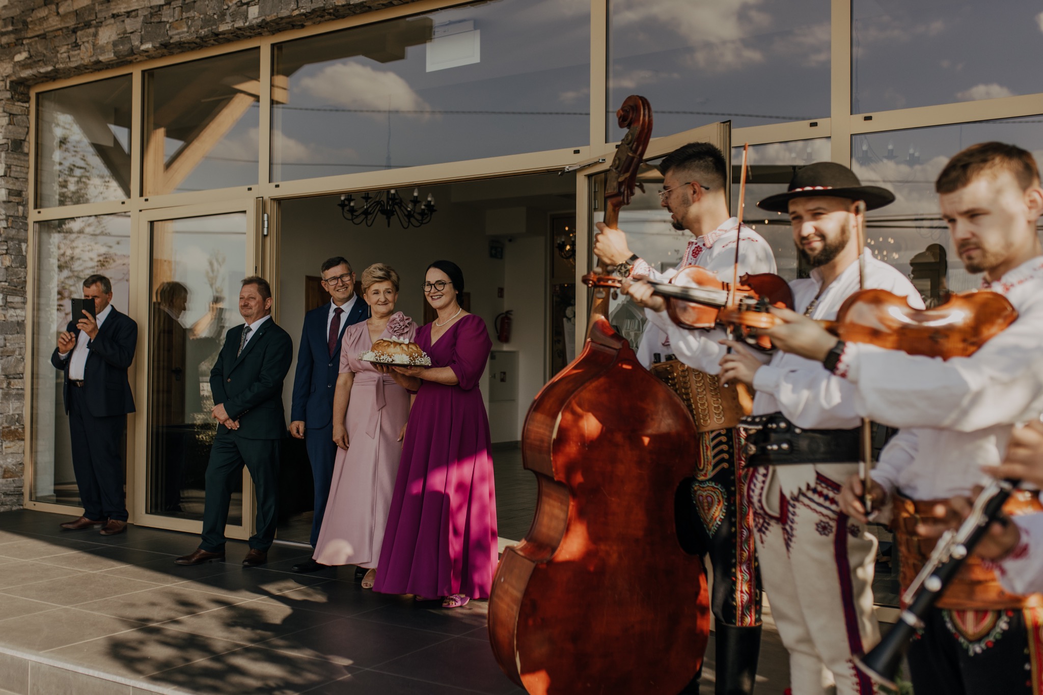 Zespół w strojach ludowych gra na skrzypcach i kontrabasie podczas powitania gości weselnych przed budynkiem z przeszklonymi drzwiami.