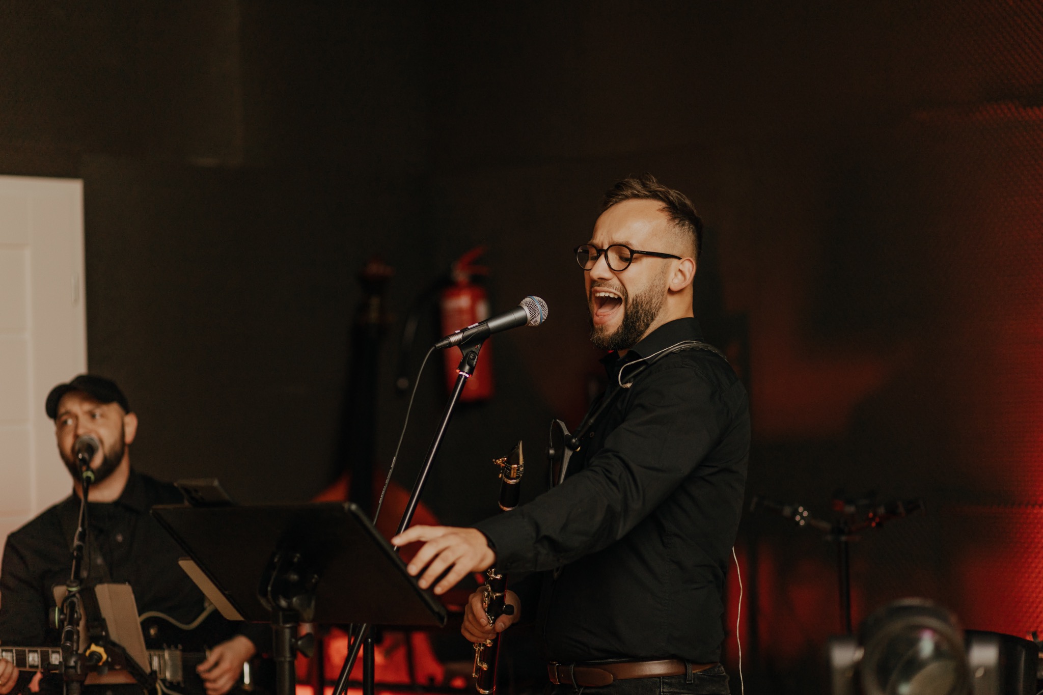 Wokalista z saksofonem śpiewa do mikrofonu podczas występu zespołu muzycznego z Nowego Sącza. Drugi muzyk gra na gitarze w tle, scena oświetlona czerwonym światłem.