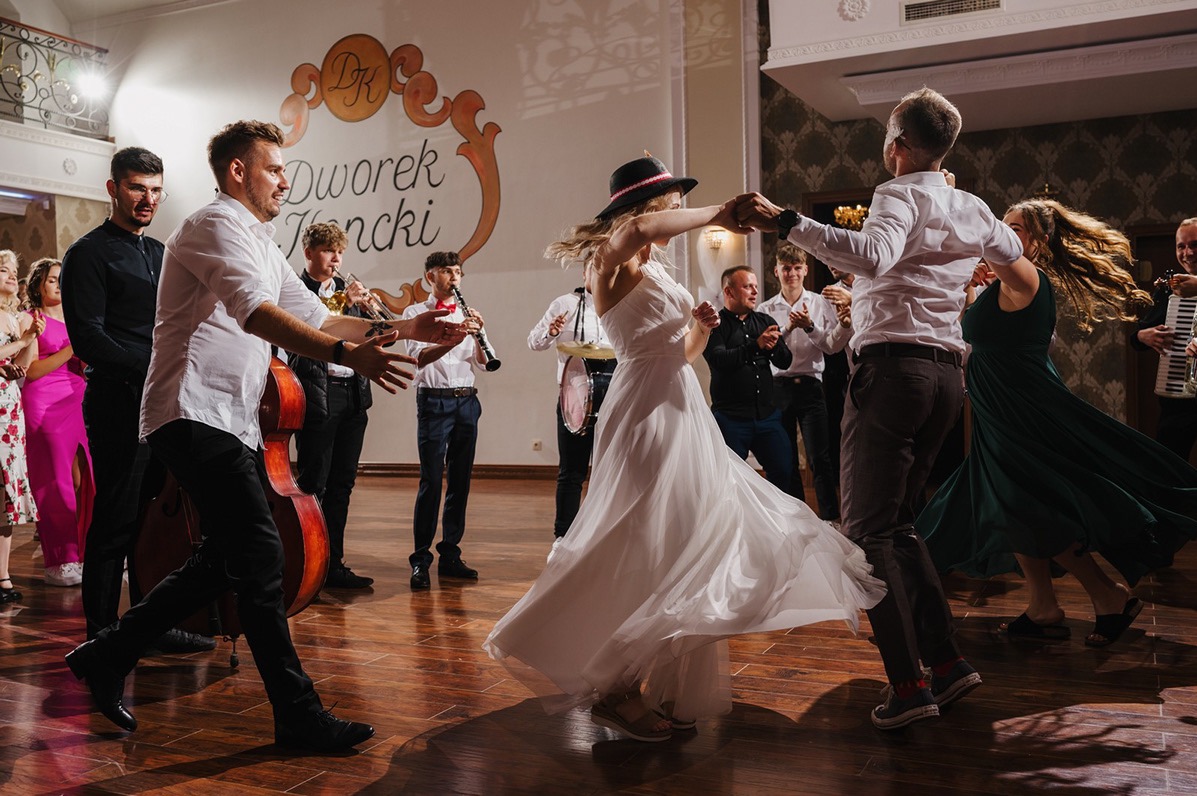 Zespół muzyczny grający na weselu, para młoda tańczy na parkiecie, w tle goście i dekoracja sali weselnej z napisem 'Dworek Jeziorki'.