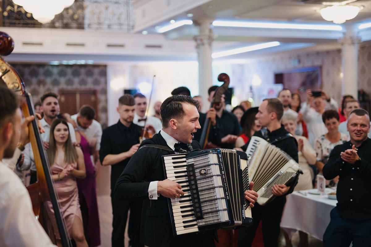 Zespół muzyczny grający na żywo na akordeonach i kontrabasach podczas wesela, z uśmiechniętymi gośćmi w tle, w eleganckiej sali bankietowej.