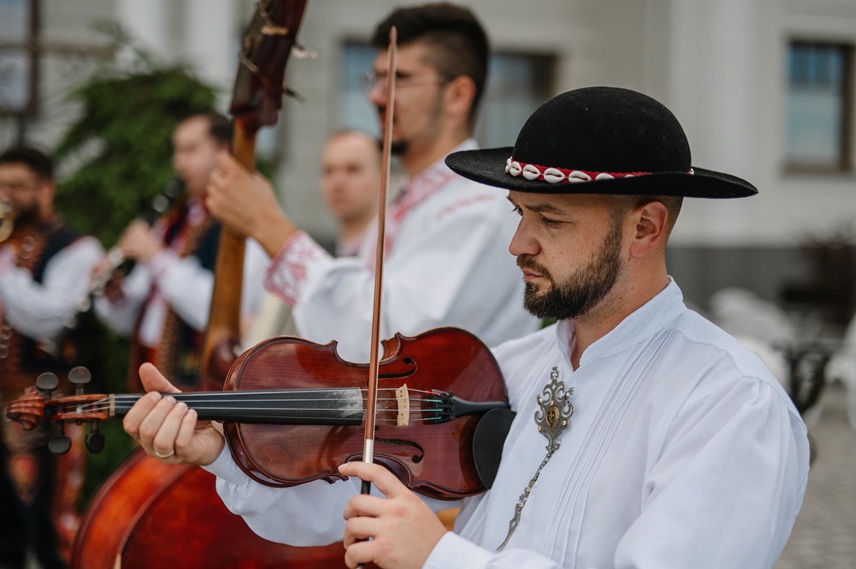 Zespół muzyczny w strojach ludowych, skrzypek w kapeluszu gra na skrzypcach. W tle kontrabasista i klarnecista. Ujęcie z bliska, skupione na skrzypku.