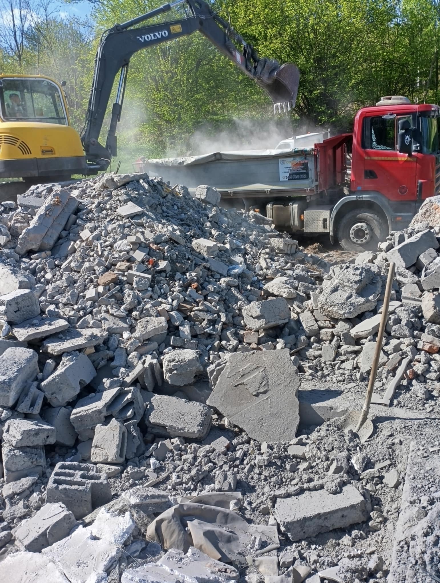 Żywiec: Żółta koparka i Volvo ładują gruz na czerwoną ciężarówkę. W tle drzewa i niebo. Szpadel wbity w gruzowisko na pierwszym planie.