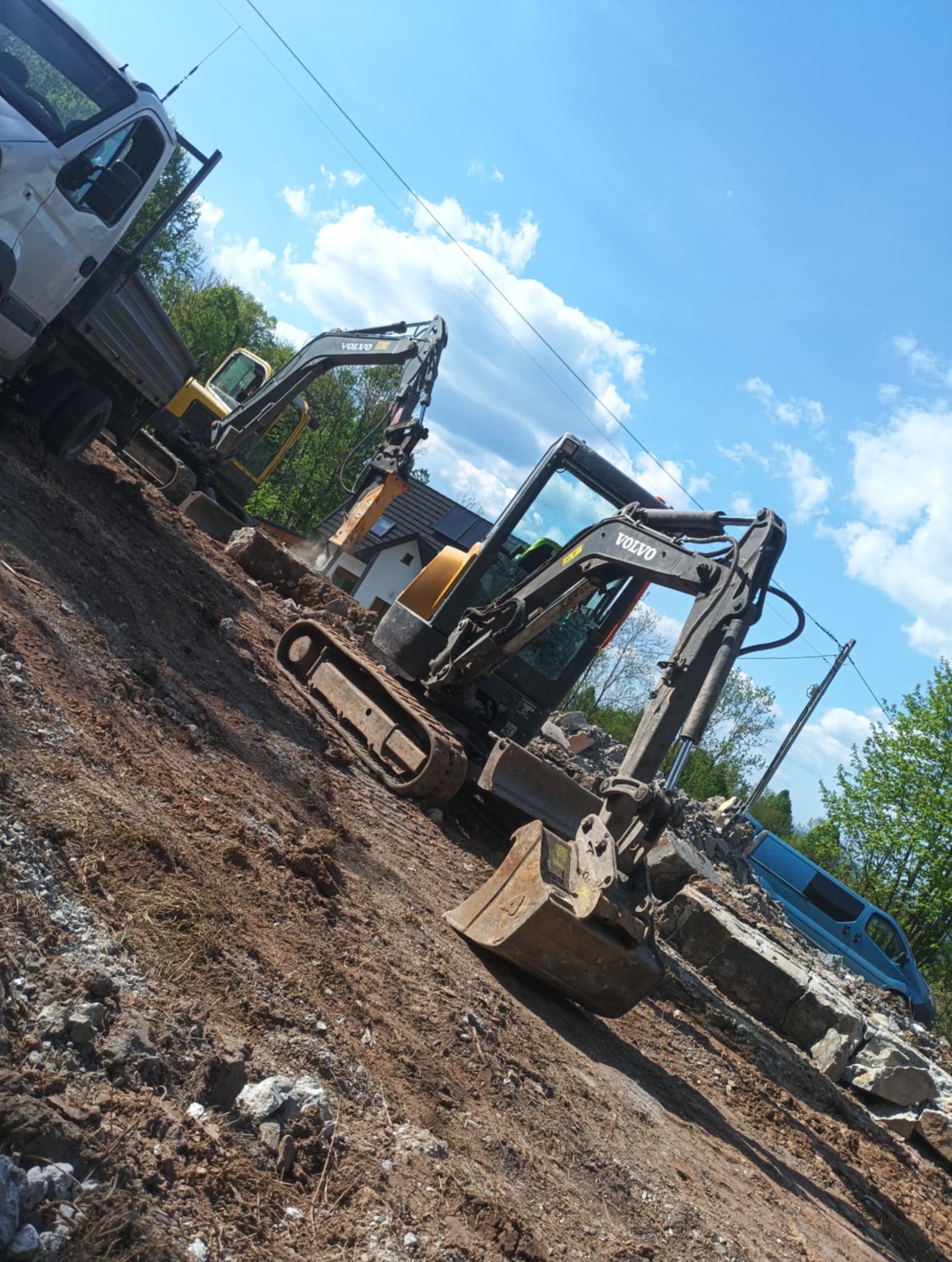 Dwie koparki Volvo na placu budowy, jedna z młotem hydraulicznym. W tle gruz, ziemia, budynek z panelami solarnymi i dostawczy samochód.