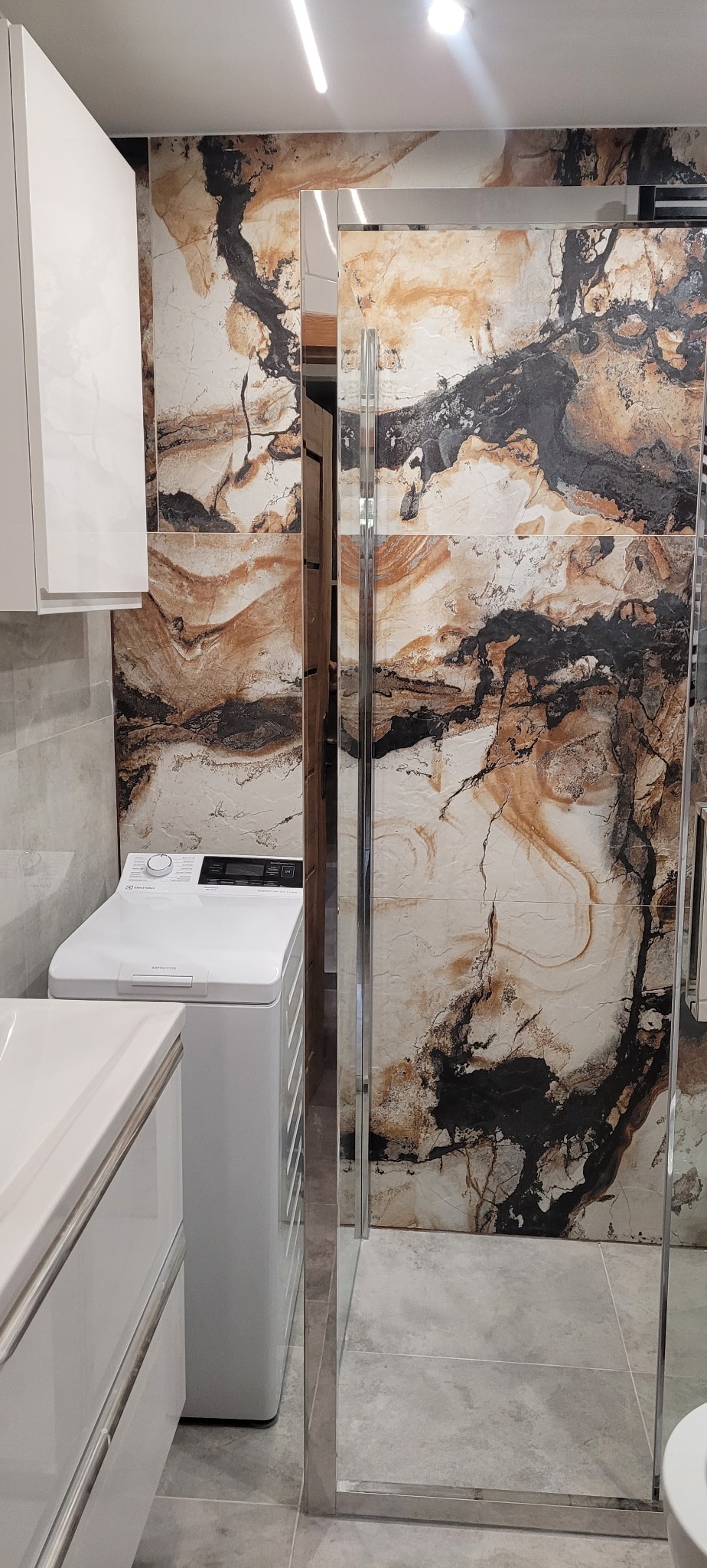 Nowoczesna łazienka z kabiną prysznicową typu walk-in, wykończona płytkami imitującymi marmur w odcieniach bieli, brązu i czerni, z pralką Electrolux i białymi szafkami.