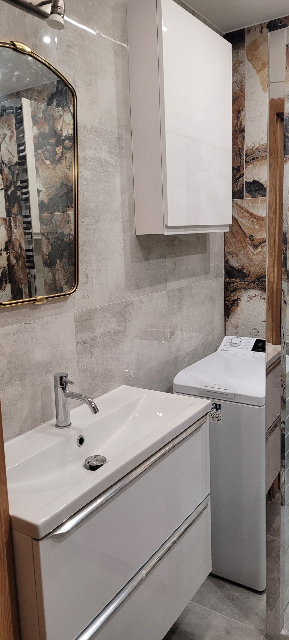 Umywalka z szafką w nowoczesnej łazience, obok pralka Electrolux i szafka wisząca, ściany wyłożone płytkami imitującymi kamień.