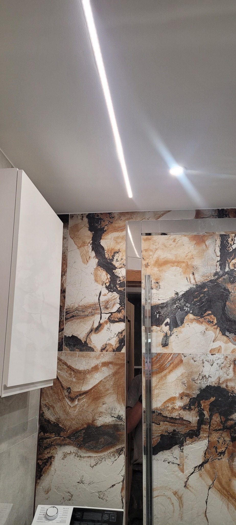 Ułożone płytki imitujące marmur na ścianie łazienki, widoczna biała szafka i oświetlenie LED na suficie, fragment pralki.