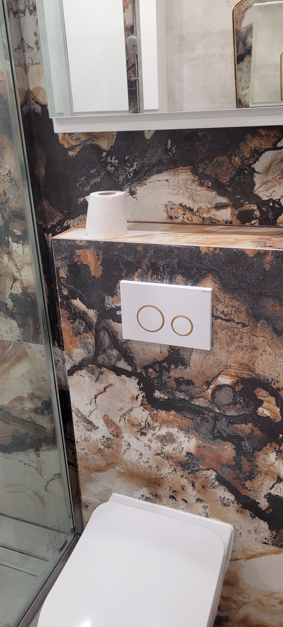 Nowoczesna toaleta wisząca z podtynkowym systemem spłukiwania z dwoma złotymi przyciskami na tle ściany wykończonej płytkami imitującymi marmur w odcieniach brązu, czerni i bieli, obok rolka...