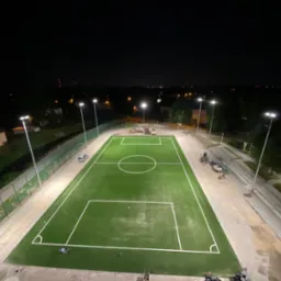 Oświetlony nocą, widok z góry na nowo wybudowane boisko z zieloną sztuczną nawierzchnią, białe linie boiska i słupy oświetleniowe, w tle zabudowania miejskie i maszyny budowlane.