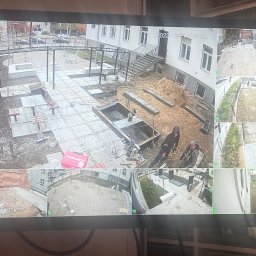 Elektron Paweł Baszak - Monitoring placu budowy z widokiem na prace ziemne, układanie kostki brukowej i budowę elementów małej architektury, widoczne osoby wykonujące prace. Data i godzina na ekranie monitoringu.