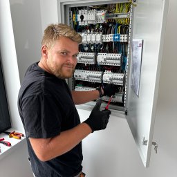 Elektron Paweł Baszak - Uśmiechnięty elektryk w czarnych rękawicach, używający czerwonego śrubokrętu do pracy w rozdzielnicy elektrycznej z otwartymi drzwiami, widoczny schemat na wewnętrznej stronie drzwi