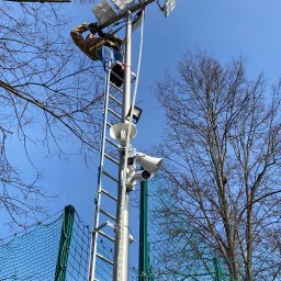 Elektron Paweł Baszak - Pracownik na drabinie montuje lub serwisuje reflektory i głośniki na wysokim maszcie oświetleniowym, widoczne drzewa i siatka ogrodzeniowa w tle.