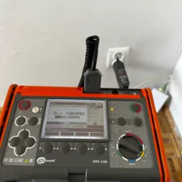Miernik Sonel MPI-530 w trakcie pomiarów elektrycznych, widoczny ekran z wynikami, gniazdko z podłączonym przewodem pomiarowym. Urządzenie na tle ściany i podłogi.
